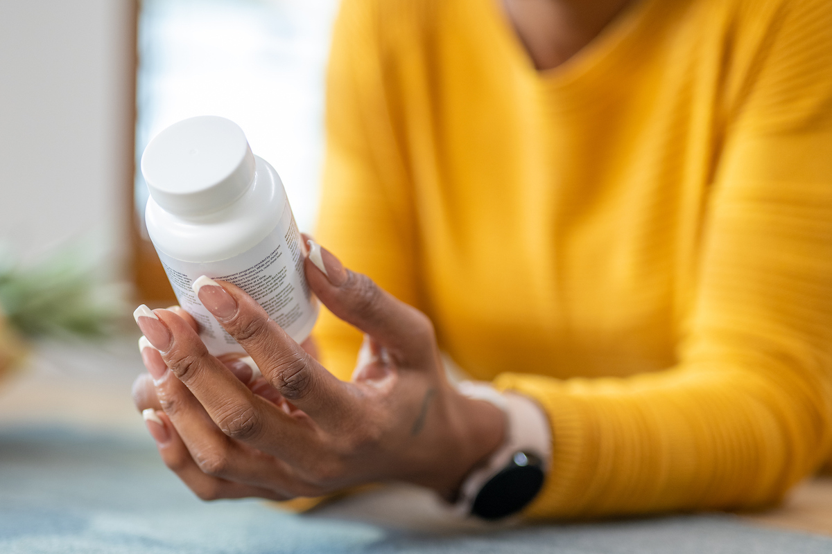 6 thành phần cần lưu ý khi dùng vitamin tổng hợp- Ảnh 1. Cận cảnh một người phụ nữ mặc áo len màu vàng cầm một viên thuốc, vitamin hoặc chai thực phẩm bổ sung và đọc thành phần