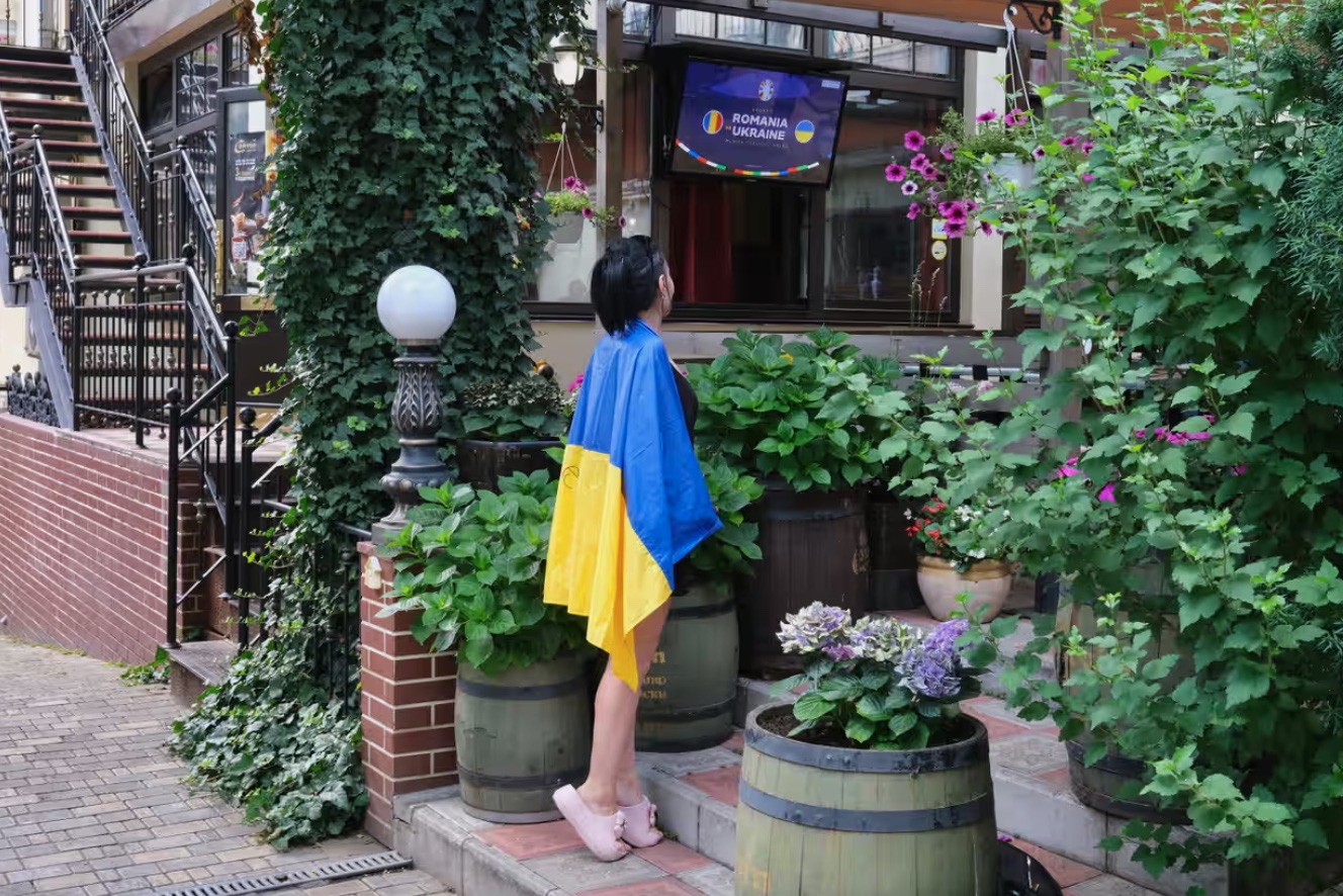 Mặc kệ chiến tranh, người dân Ukraine hòa vào không khí Euro 2024- Ảnh 9. Một cô gái Ukraine đang khoác quốc kỳ lên người và háo hức chờ đợi trận đấu giữa Ukraine và Romania. Trên quốc kỳ, cô viết to cụm từ “Kherson” - tên một thành phố ở Ukraine và cũng là quê hương của cô. Tuy nhiên, cô gái này đã phải ly hương trước các cuộc không kích của Nga - Ảnh: RICHARD MORGAN